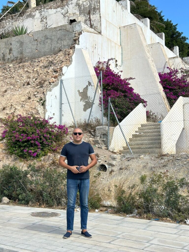 Hombre de pie frente a una pared deteriorada con flores moradas.