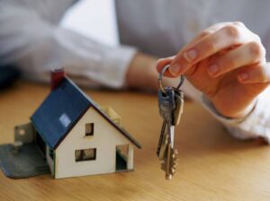 Persona sosteniendo llaves frente a una casa en miniatura