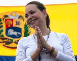 María Corina Machado sonriendo con las manos en posición de oración frente a una bandera de Venezuela.