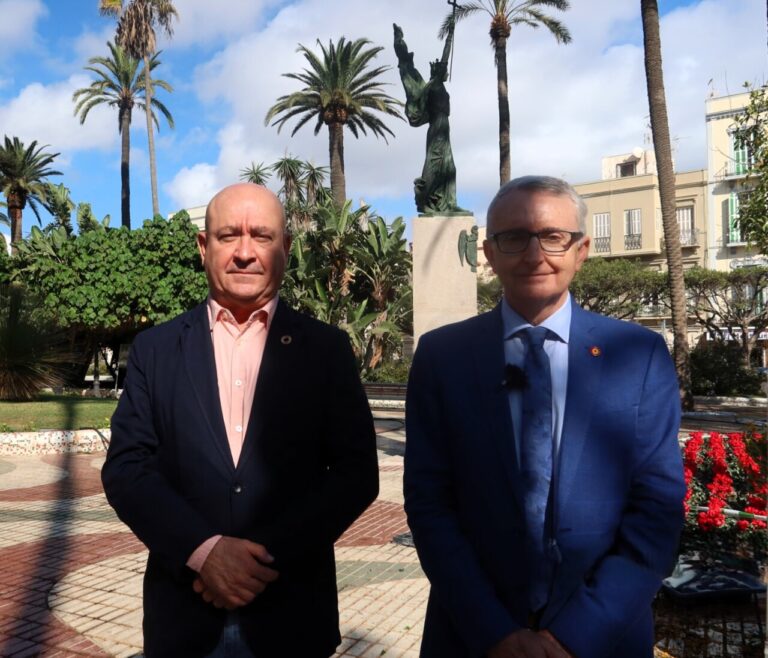 Dos hombres posando en un parque con palmeras y una estatua detrás.