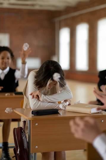 Estudiante triste en clase mientras otros lanzan papeles.