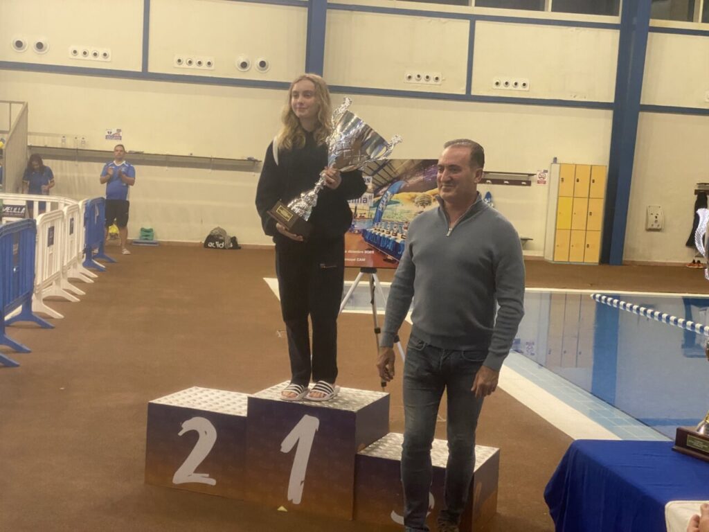 Ganadora del Trofeo de Natación Ciudad de Melilla sosteniendo el trofeo en el podio