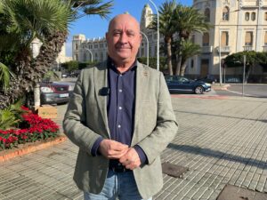 Hombre con chaqueta gris posando en una calle con palmeras.