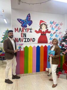 Dos personas sosteniendo libros frente a un mural navideño colorido.