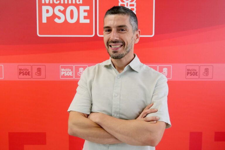 Persona sonriendo con brazos cruzados frente a un fondo rojo del PSOE
