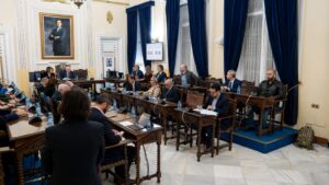 Reunión en un salón con varias personas sentadas y un retrato en la pared.