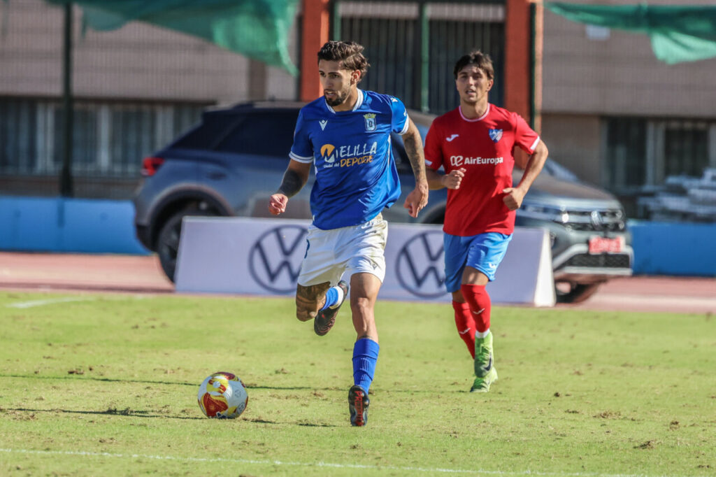 Jugador de la U.D. Melilla corriendo con el balón durante un partido de fútbol.