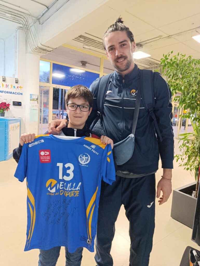 Un niño sostiene una camiseta del Club Voleibol Melilla junto a un jugador.
