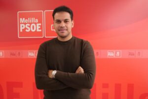 Persona de pie con brazos cruzados frente a un fondo rojo con logos del PSOE.