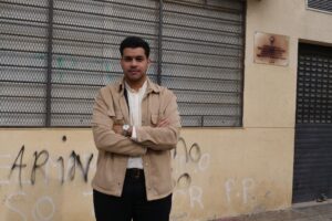 Hombre de pie frente a una pared con graffiti y rejas metálicas.