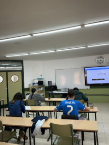 Estudiantes en un aula realizando un examen con un reloj visible.
