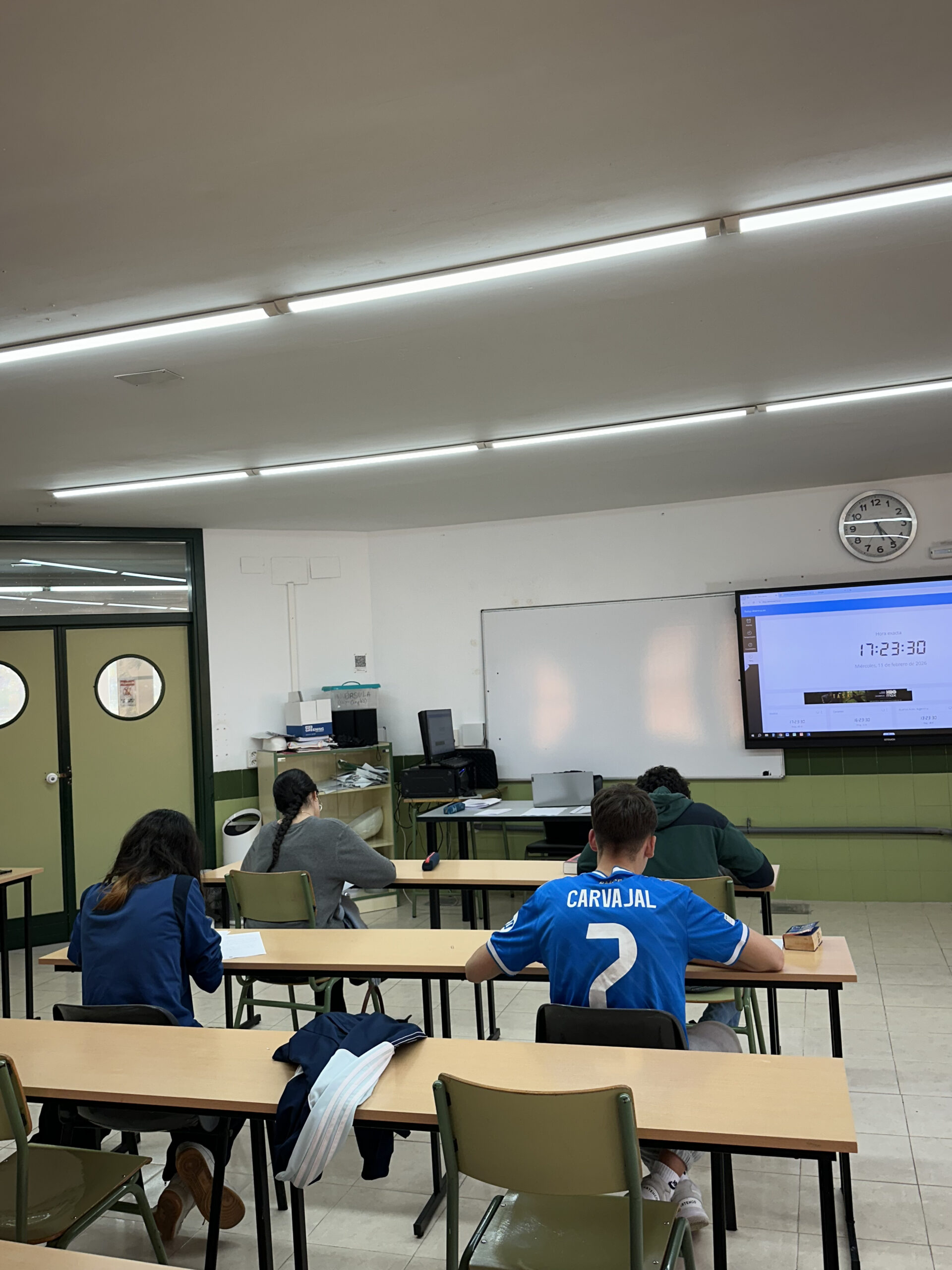 Estudiantes en un aula realizando un examen con un reloj visible.