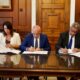 Tres personas firmando un convenio en una mesa de madera