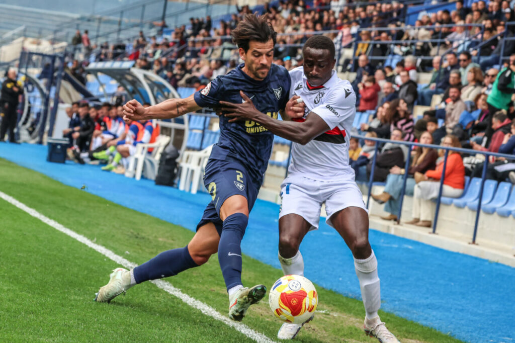 Jugadores de U.D. Melilla y UCAM Murcia compiten por el balón en un partido de fútbol.