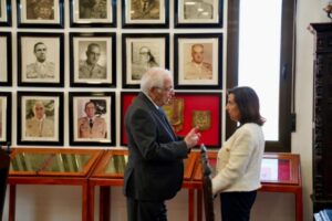 Dos personas conversando en un entorno institucional con retratos en la pared.