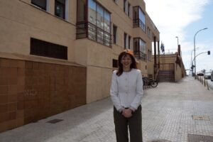 Mujer con cabello castaño y chaqueta blanca sonríe frente a un edificio.