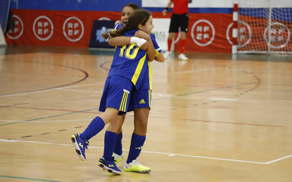 Two young female teammates in blue and yellow uniforms embrace in celebration on an indoor futsal court, with a goal net in the background.