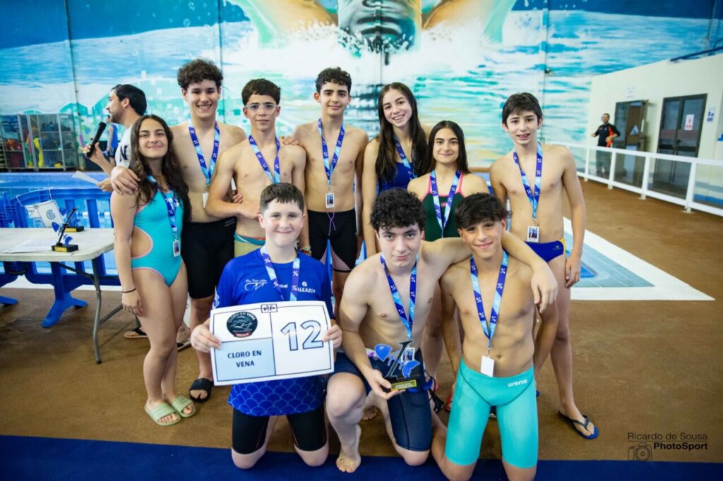 Group of ten young swimmers with medals posing by an indoor pool after a race, two in front hold a certificate and a trophy.