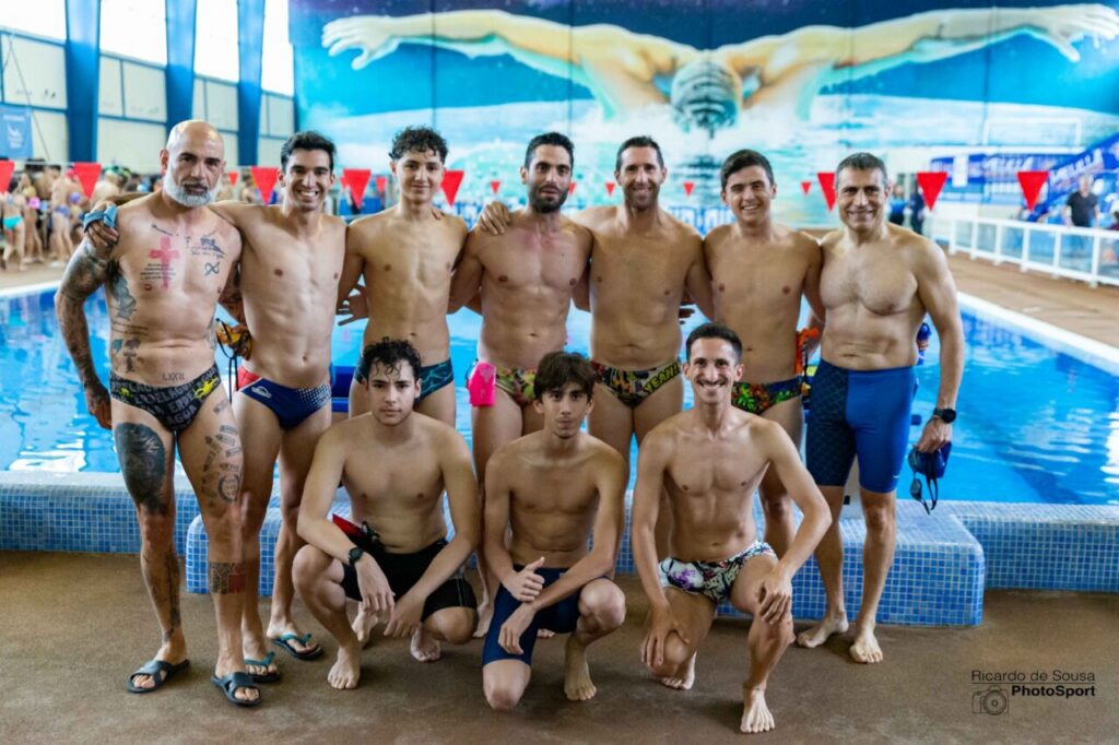 Group of ten male swimmers posing together by an indoor pool, some kneeling in front and others standing, wearing swim briefs or trunks with a mural of a swimmer on the wall behind them.