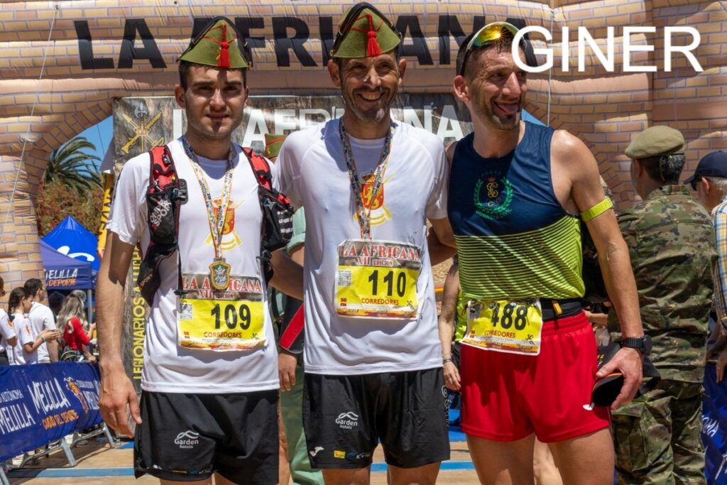 Three male runners pose with medals at the La Africana finish line; bib numbers 109, 110 and 488, with banners and spectators behind them.