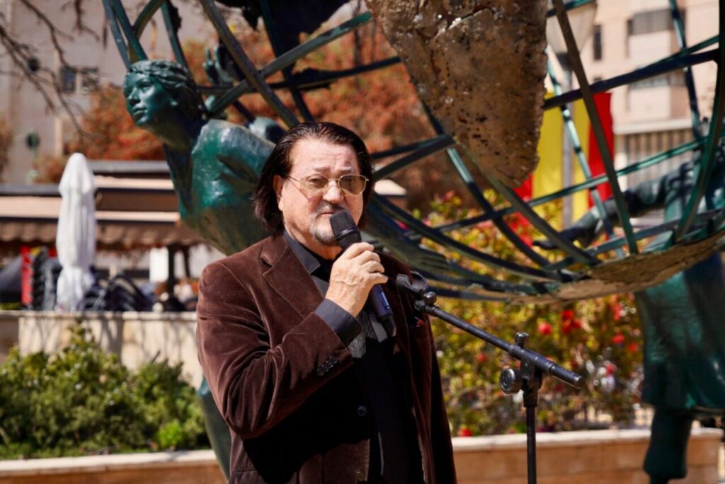 Man in a brown blazer microphone in hand, speaking outdoors at a public event with a metal sculpture behind him