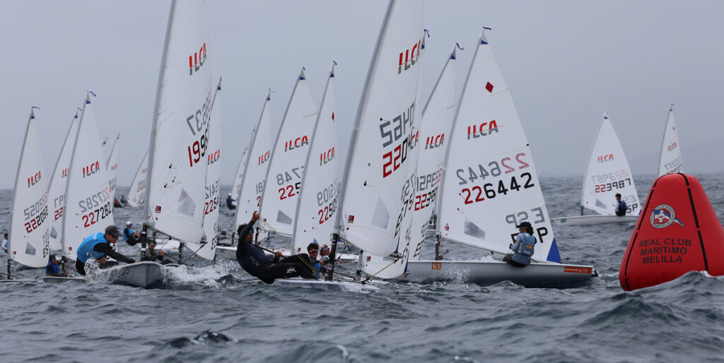 Dinghy sailing race with many small boats heeling in choppy water near a red race buoy marked Real Club Marítimo Melilla in the background.