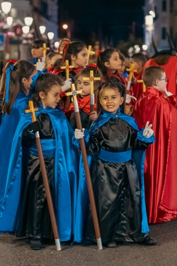 Niños cofrades con capas azules y rojas sosteniendo cruces durante la procesión.