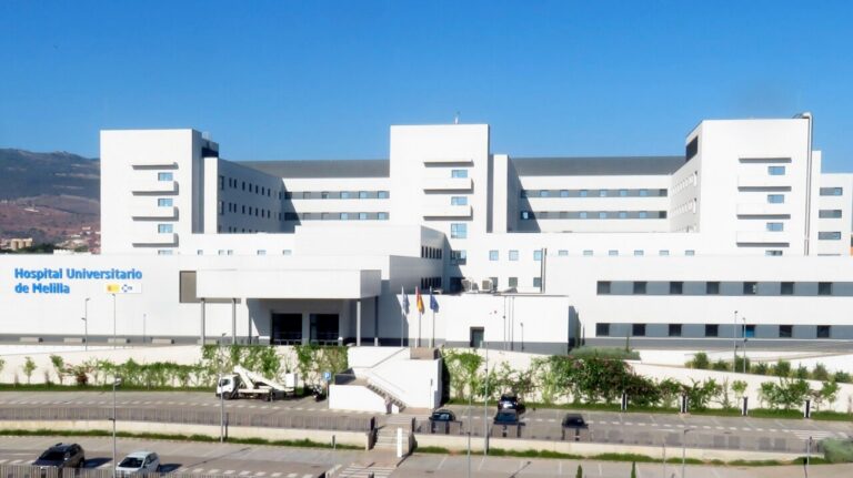 Modern white hospital building with the sign 'Hospital Universitario de Melilla' under a clear blue sky.