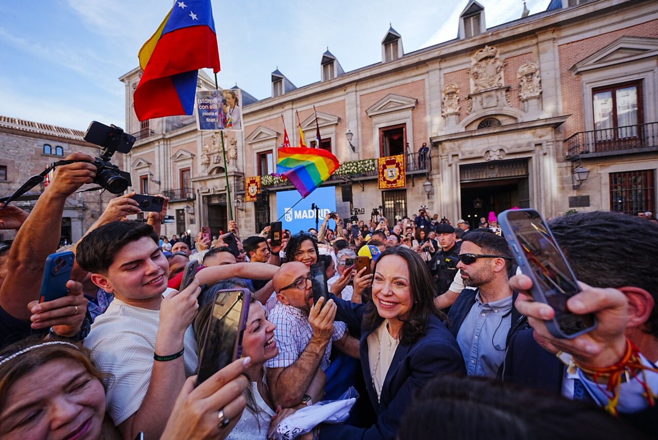 Crowd of supporters crowded around a smiling woman in a dark blazer at a public event in Madrid, waving flags including rainbow and Venezuelan flags.
