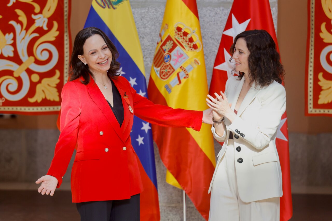 Two women in formal attire smile and shake hands at an official ceremony, with flags of Spain, Venezuela, and Turkey in the background.