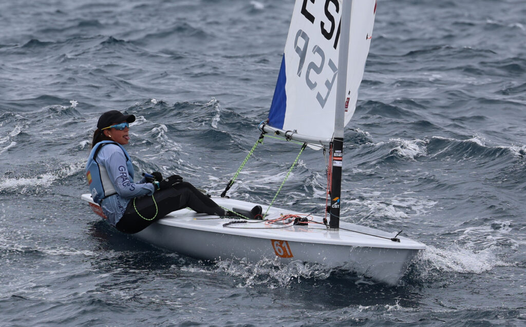 Sailor wearing cap and life vest steers a small sailboat through rough waves, number 61 on the hull, using a bright-blue sail.