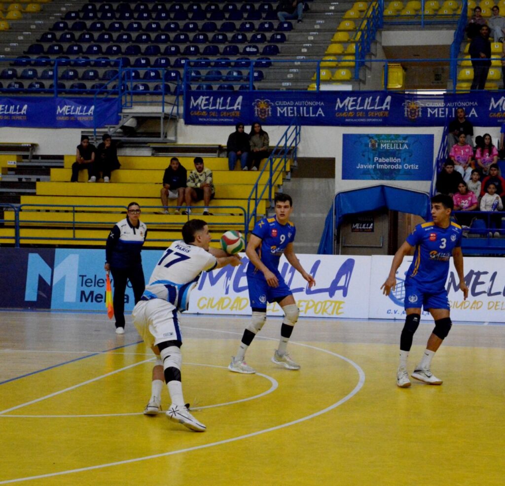 Futsal match on a yellow court; white-uniformed player #17 passes a ball to two blue-uniform players as spectators watch from yellow and blue stands with Melilla banners