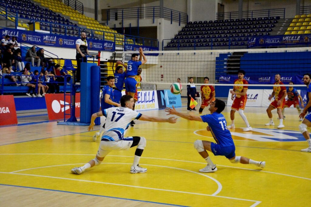 Two volleyball players dive to reach the ball near the net as blue and white teams clash on a bright yellow court, with spectators in the stands.
