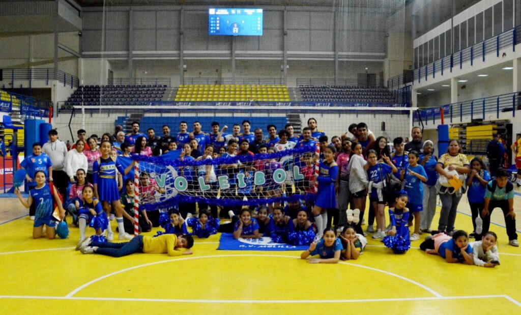Group photo of children, teens, and adults in blue uniforms posing on an indoor volleyball court with a net and banner reading VOLLEIBOL behind them.