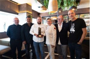 Six people in a restaurant pose for a group photo; a man in a white chef coat holds a trophy at center