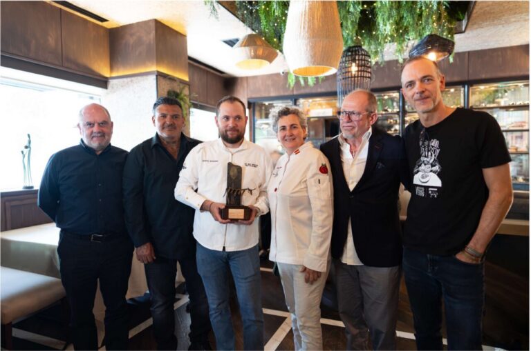 Six people in a restaurant pose for a group photo; a man in a white chef coat holds a trophy at center