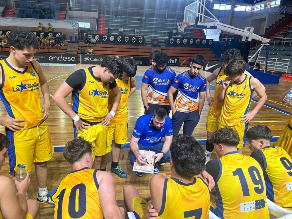 Basketball team in yellow uniforms gathers around a coach drawing plays on a clipboard on a wooden court during a timeout in the gym.