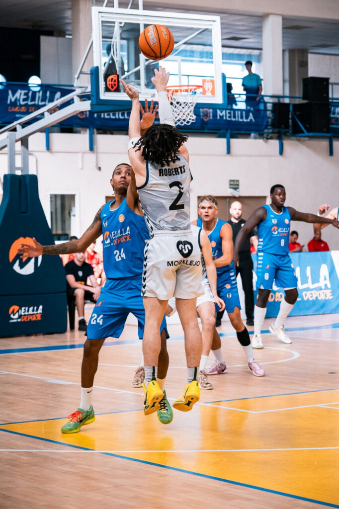 White-clad basketball player (#2, Roberts) leaps toward the hoop for a layup as blue-team defenders watch from nearby.