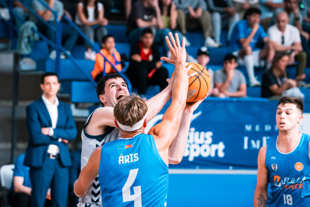 Two basketball players leap for the ball in a contested rebound as spectators watch from the stands nearby