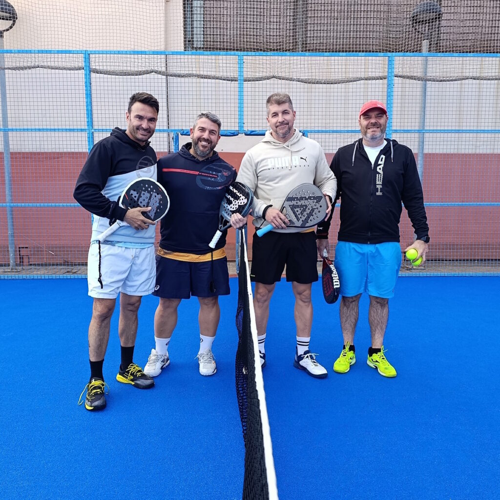 Four men pose on a blue padel court, each holding a padel racket, smiling at the camera.