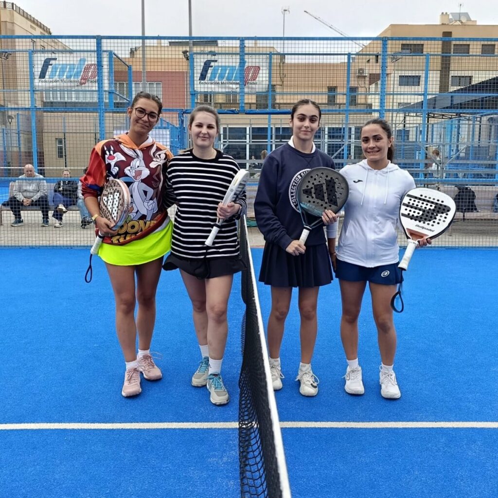 Four young women on a blue padel court pose with paddles, wearing sporty outfits.