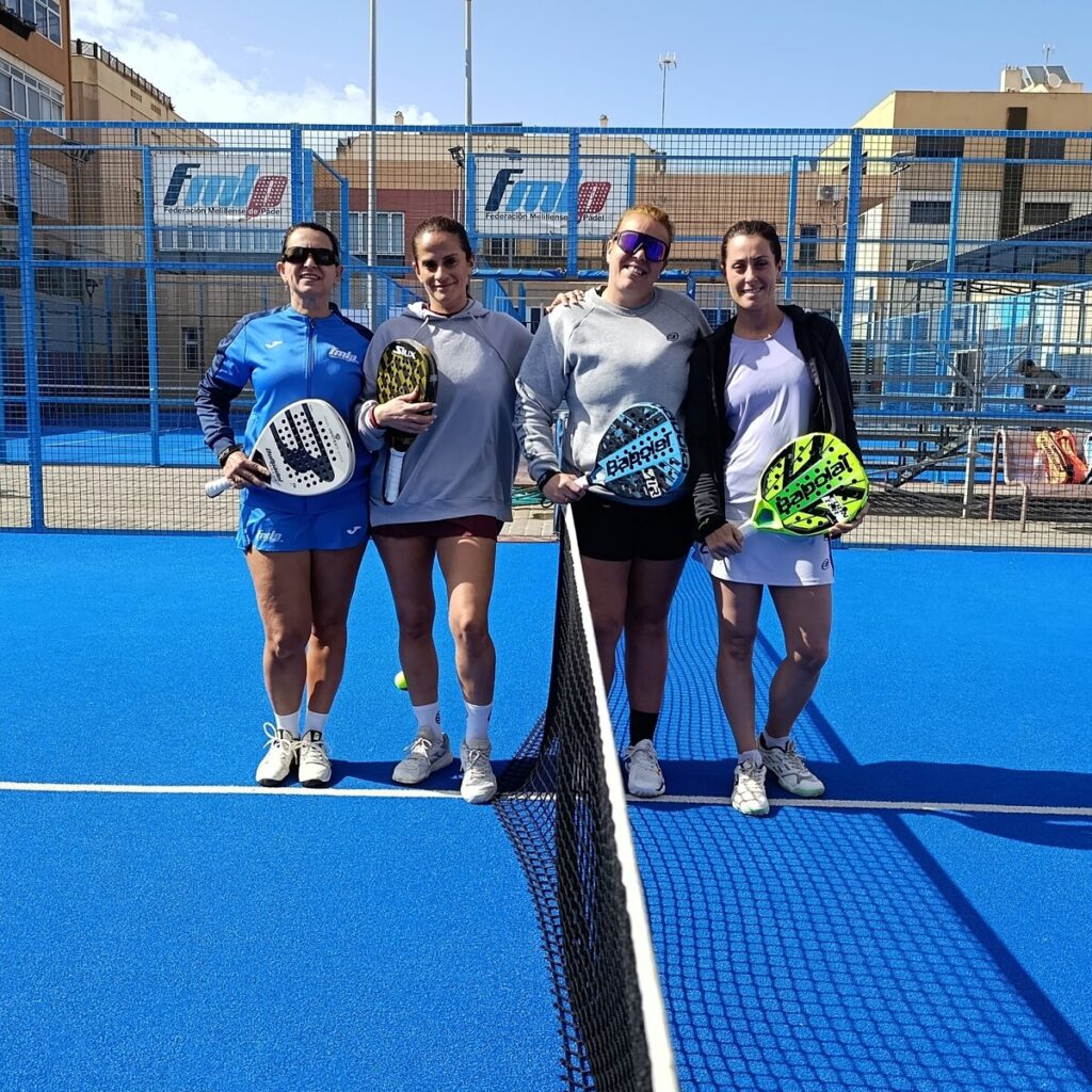 Four women stand together on a blue padel court, each holding a paddle and smiling after a game on a sunny day.