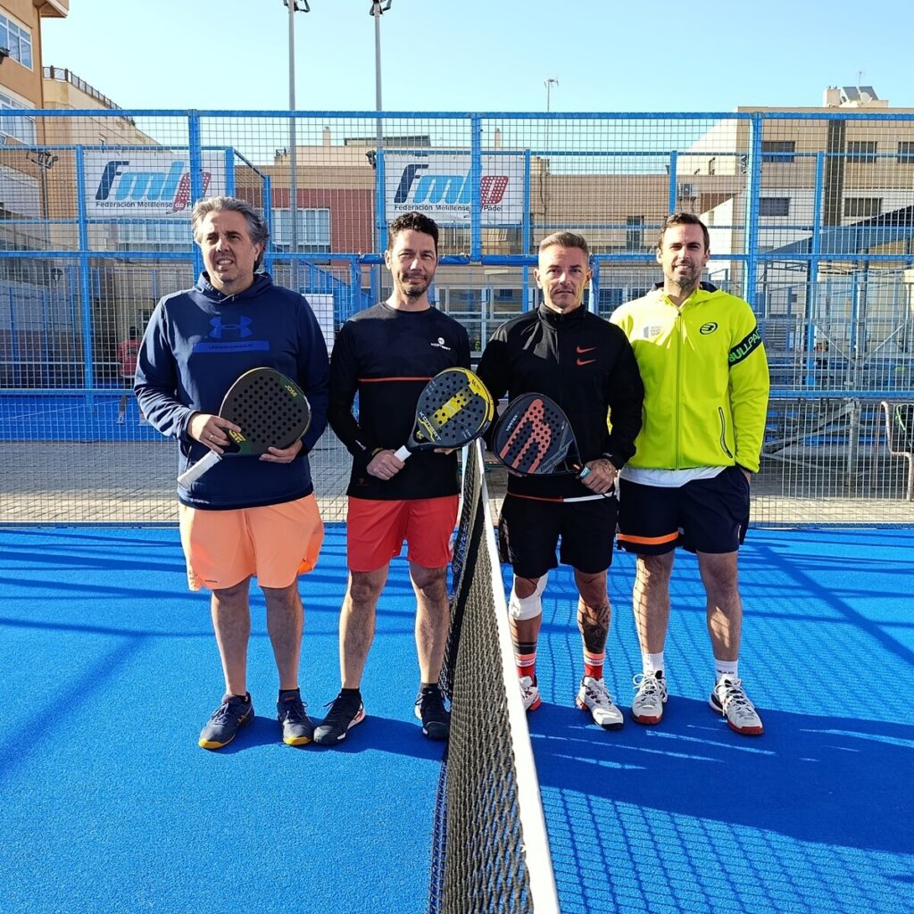 Four men standing on a blue padel court behind a net, each holding paddle racquets and smiling on a sunny day.