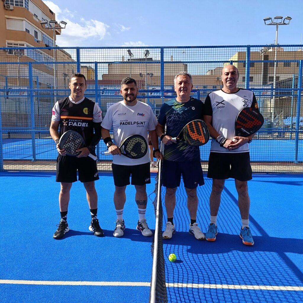 Four men stand side by side on a blue paddle tennis court, each holding a paddle and smiling toward the camera, with a net and urban buildings in the background.