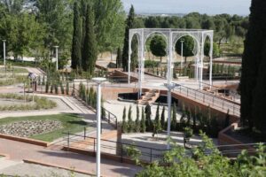 Public park with brick terraces, white metal arches, and winding paths among trees and shrubs to the center. ( informative )