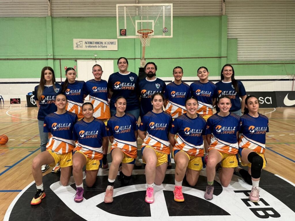 Group of female players in blue and orange jerseys posing for a team photo in an indoor basketball gym, with a coach in the center and a hoop visible in the background.