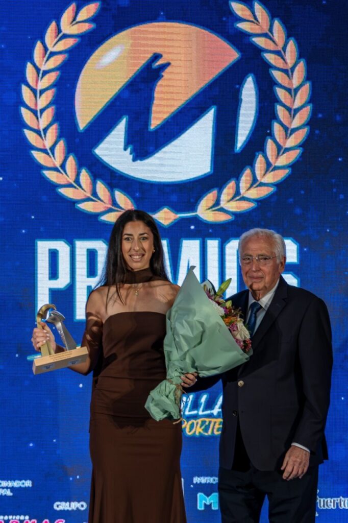 Young woman in a brown gown holding a trophy and bouquet on stage with an older man in a suit beside her,Award backdrop behind them
