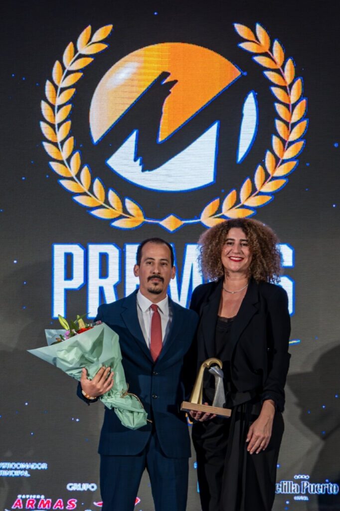 Man in a navy suit with a red tie holds a bouquet of flowers, standing beside a woman in a black blazer who holds a curved trophy; they pose on a stage with a large logo backdrop.