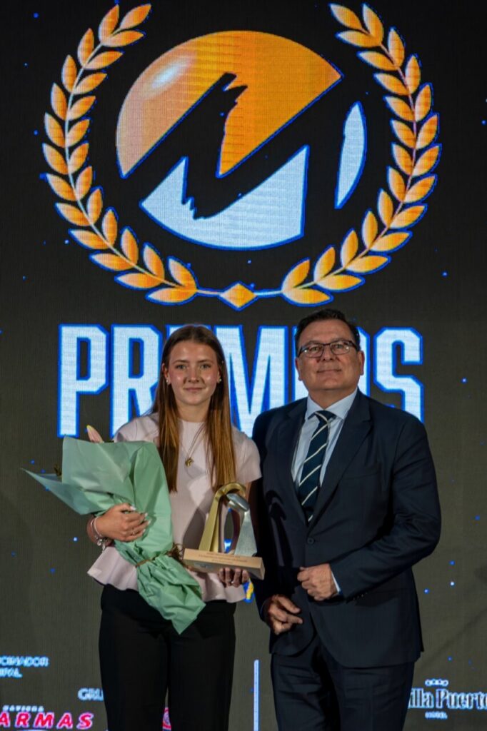 Young woman holds bouquet and trophy on stage beside a suited man during an award ceremony with a large laureled logo backdrop behind them.