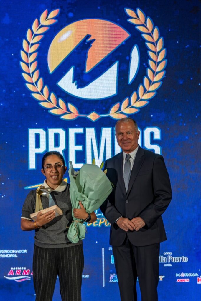 Award recipient holding a bouquet and trophy on stage, standing with a man in a suit against a large blue awards backdrop.